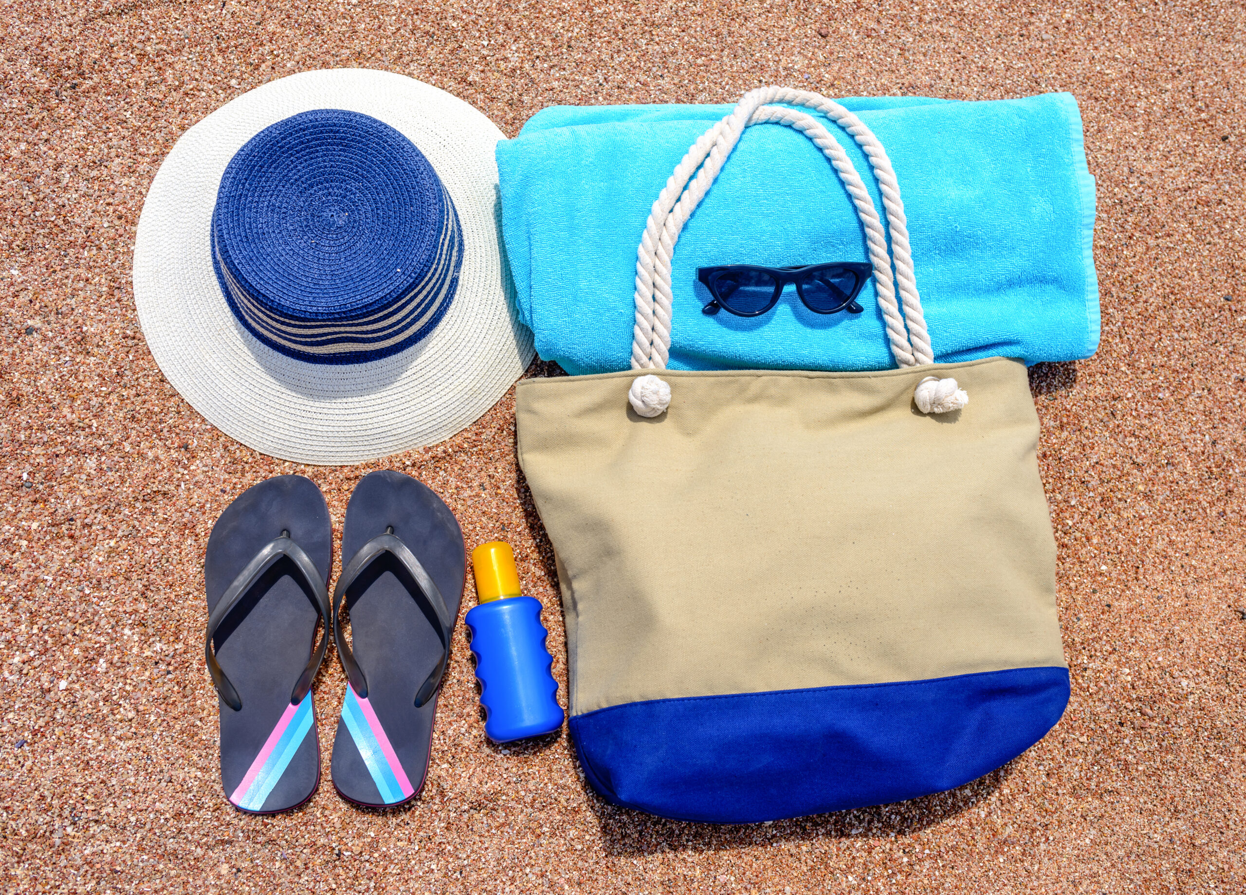 Sunhat, flip flops, towel, sunglasses, sunscreen and beach bag neatly laid out on a sandy tropical beach.