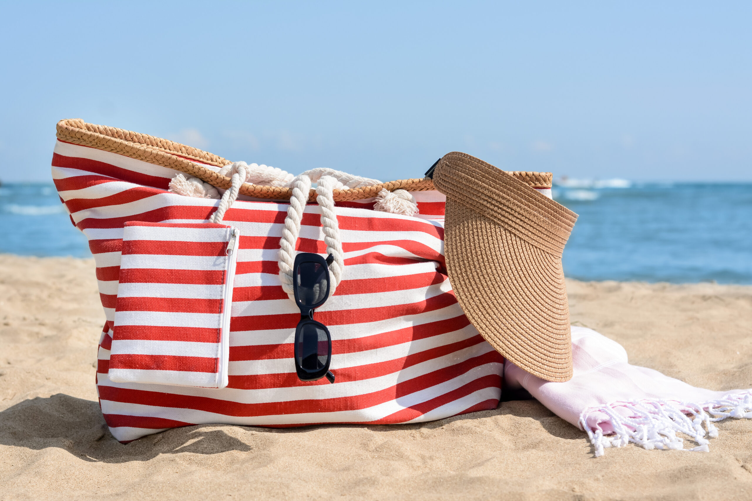 Stylish striped bag with visor cap, sunglasses and blanket on sandy beach near sea for beach side getaway.