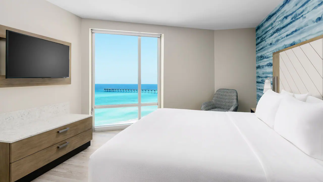 Hyatt Place guest bedroom with a white kind bed, white walls, and a big glass door overlooking the Gulf. 