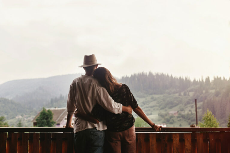 Hipster couple hugging on porch of wooden house looking at mountains in evening sunset.