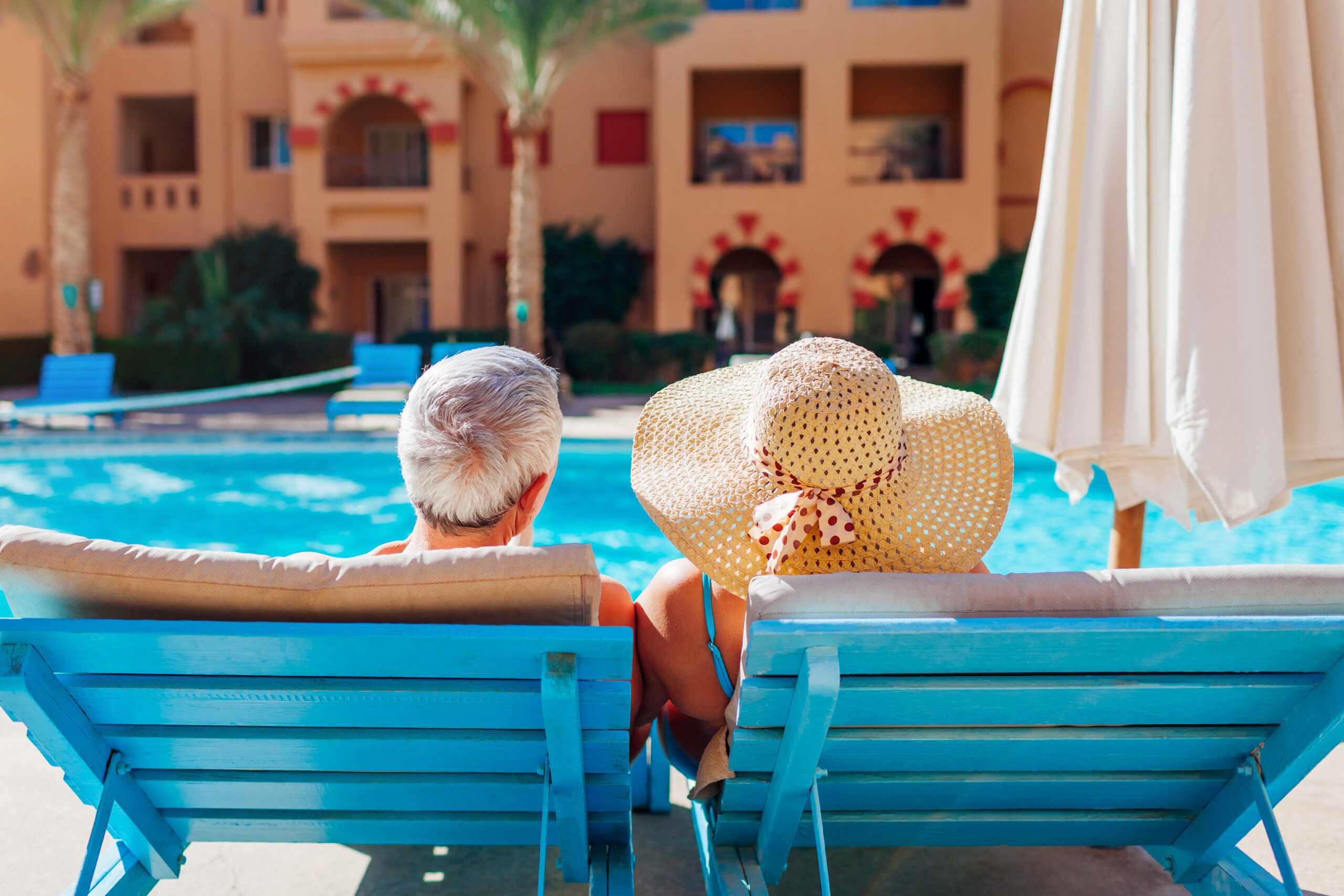 Senior couple relaxing by swimming pool lying on chaise-longues. People enjoying vacation.