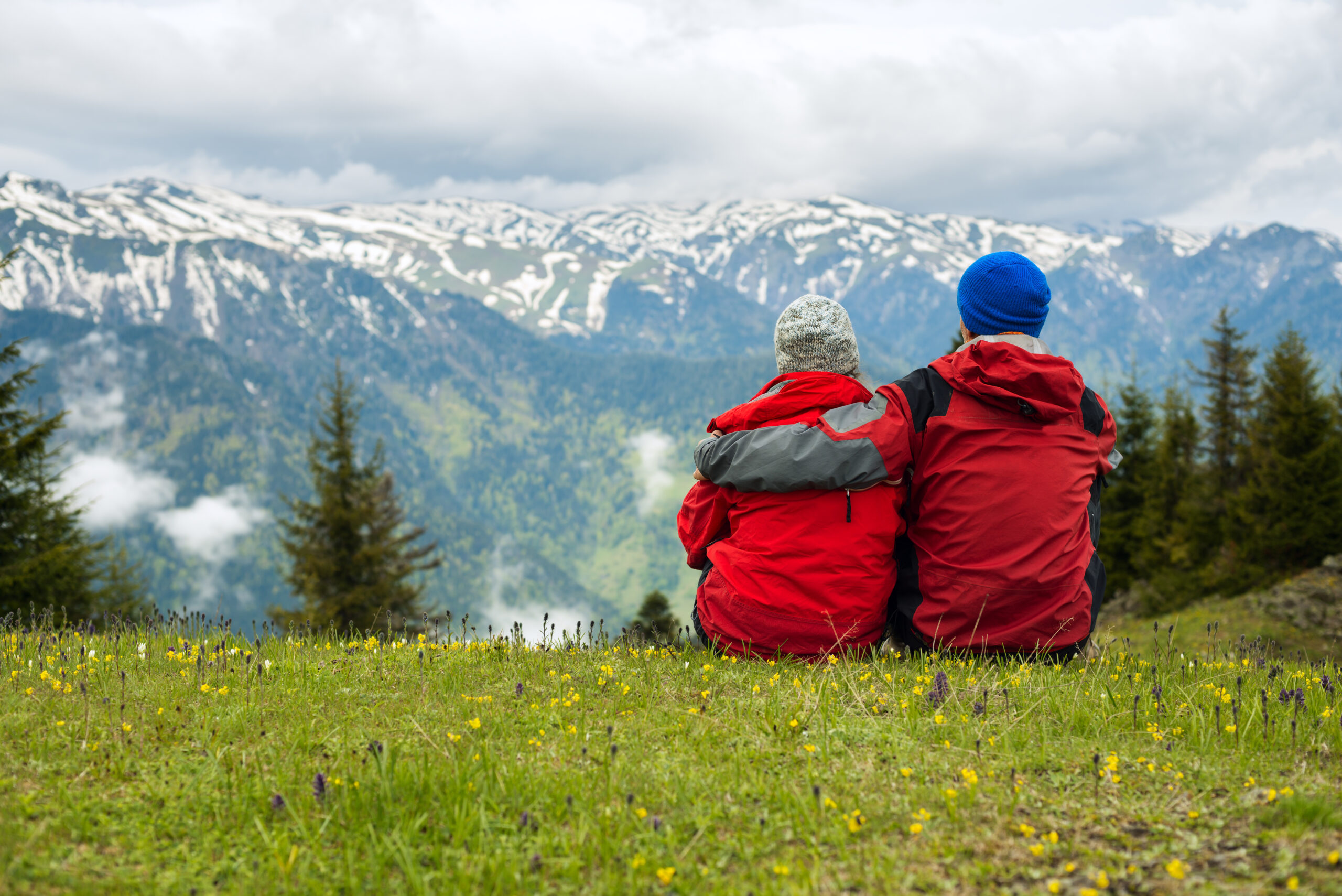 Couple of travelers sit, embracing in an alpine meadow and admiring the panorama of the mountains on the horizon.