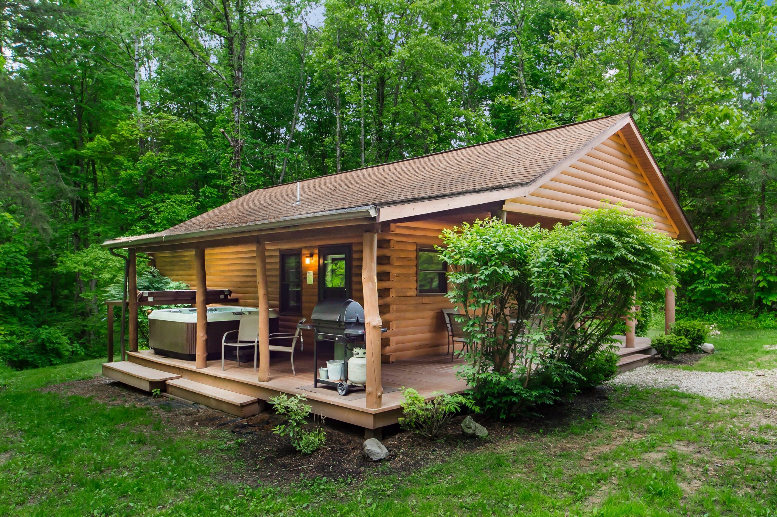 Small cabin with a hot tub and grill on front deck surrounded by trees and greenery.