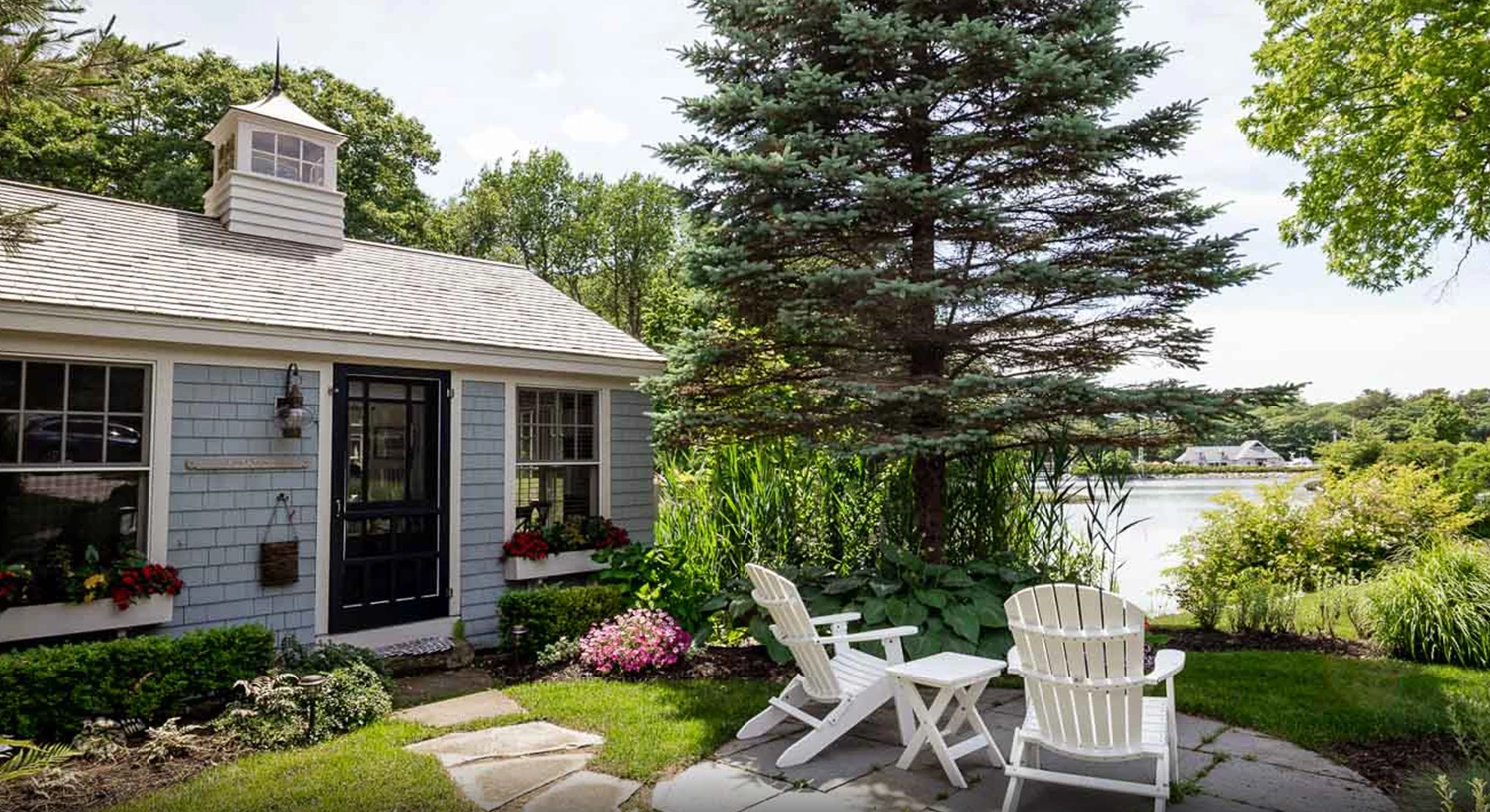 A pale blue cottage by the water with pine trees, Adirondack chairs, and blue sky. It's one of the most secluded romantic getaways in the USA!