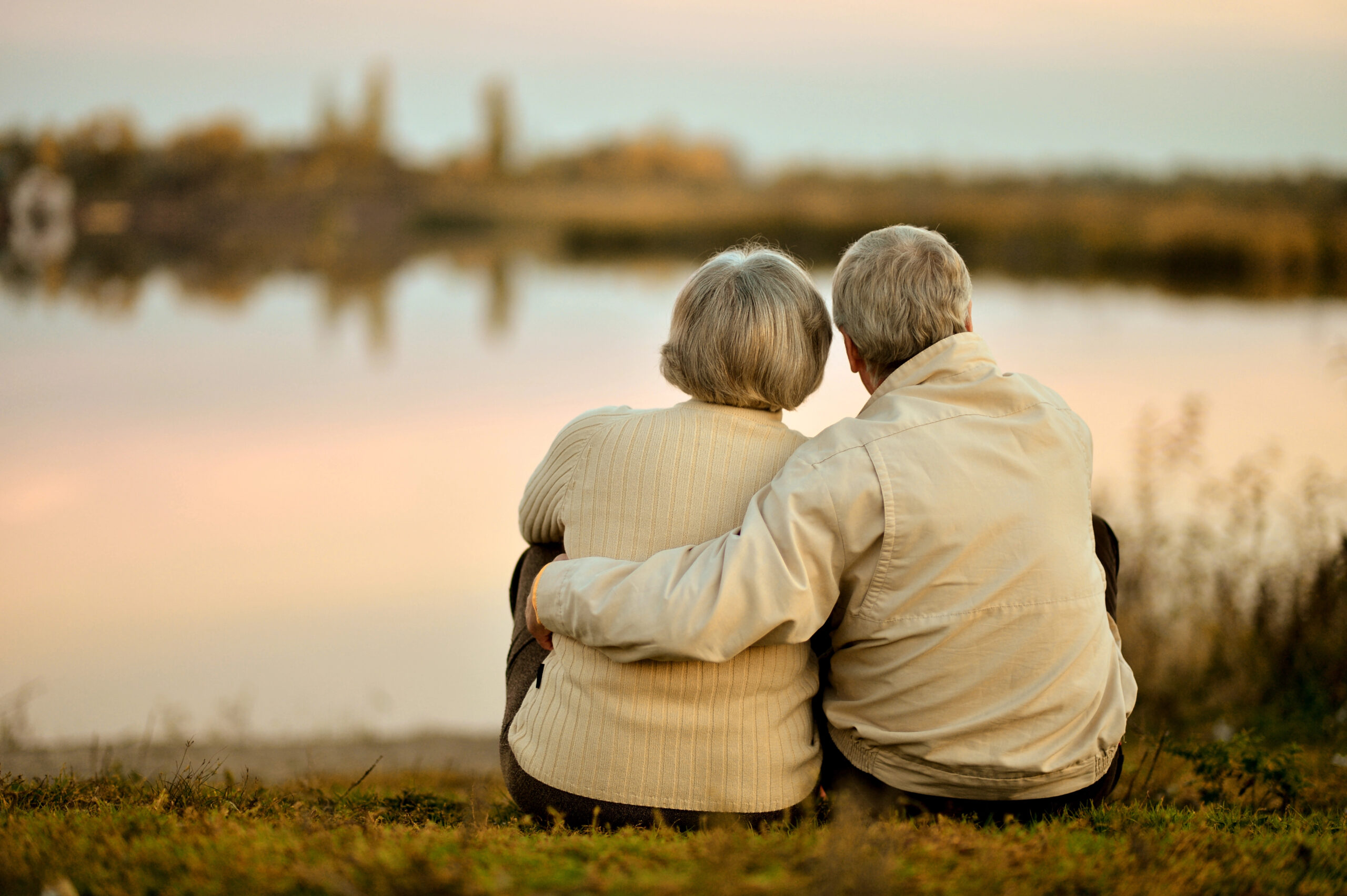 Senior couple holding each other close looking out at a lake at sunset.