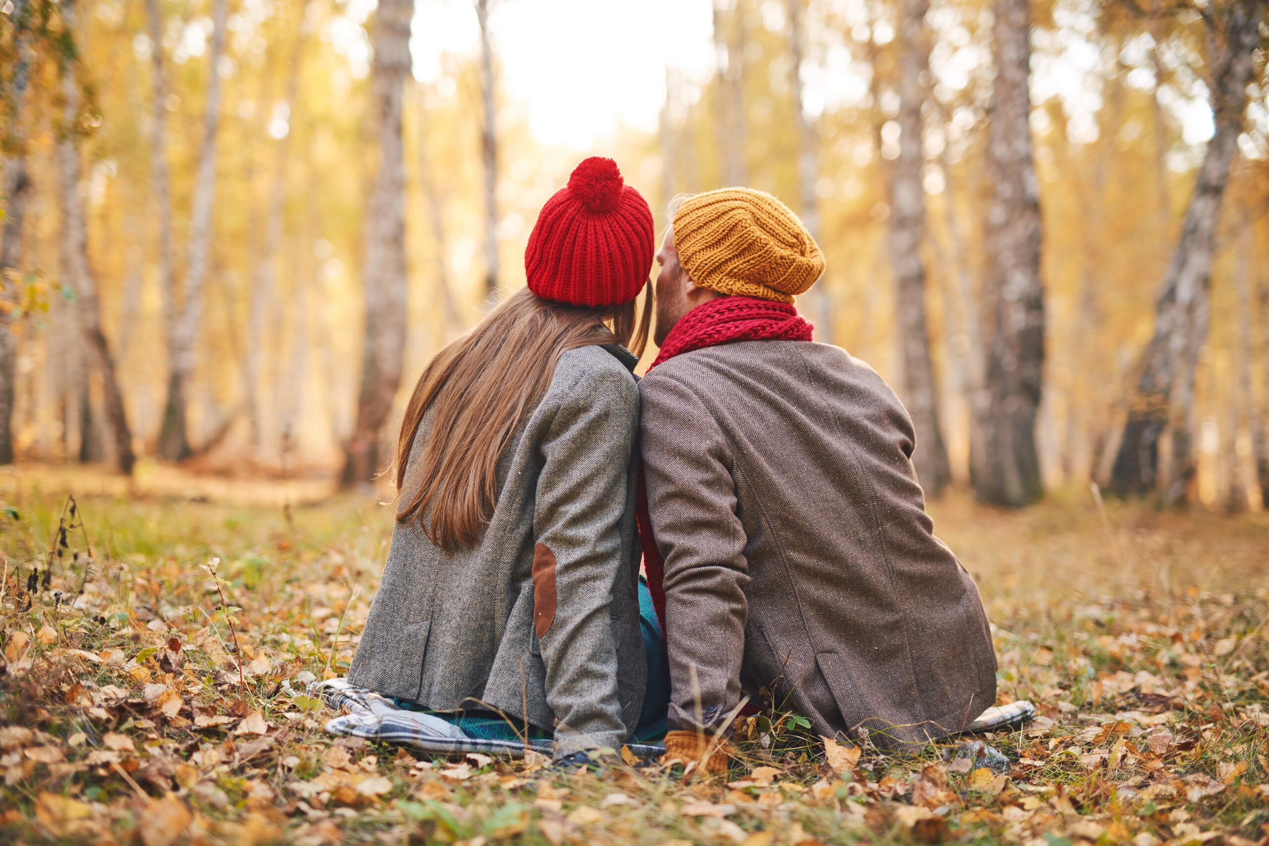 Couple sitting close in autumn woods with beanies on having the most romantic getaway in the Fall.