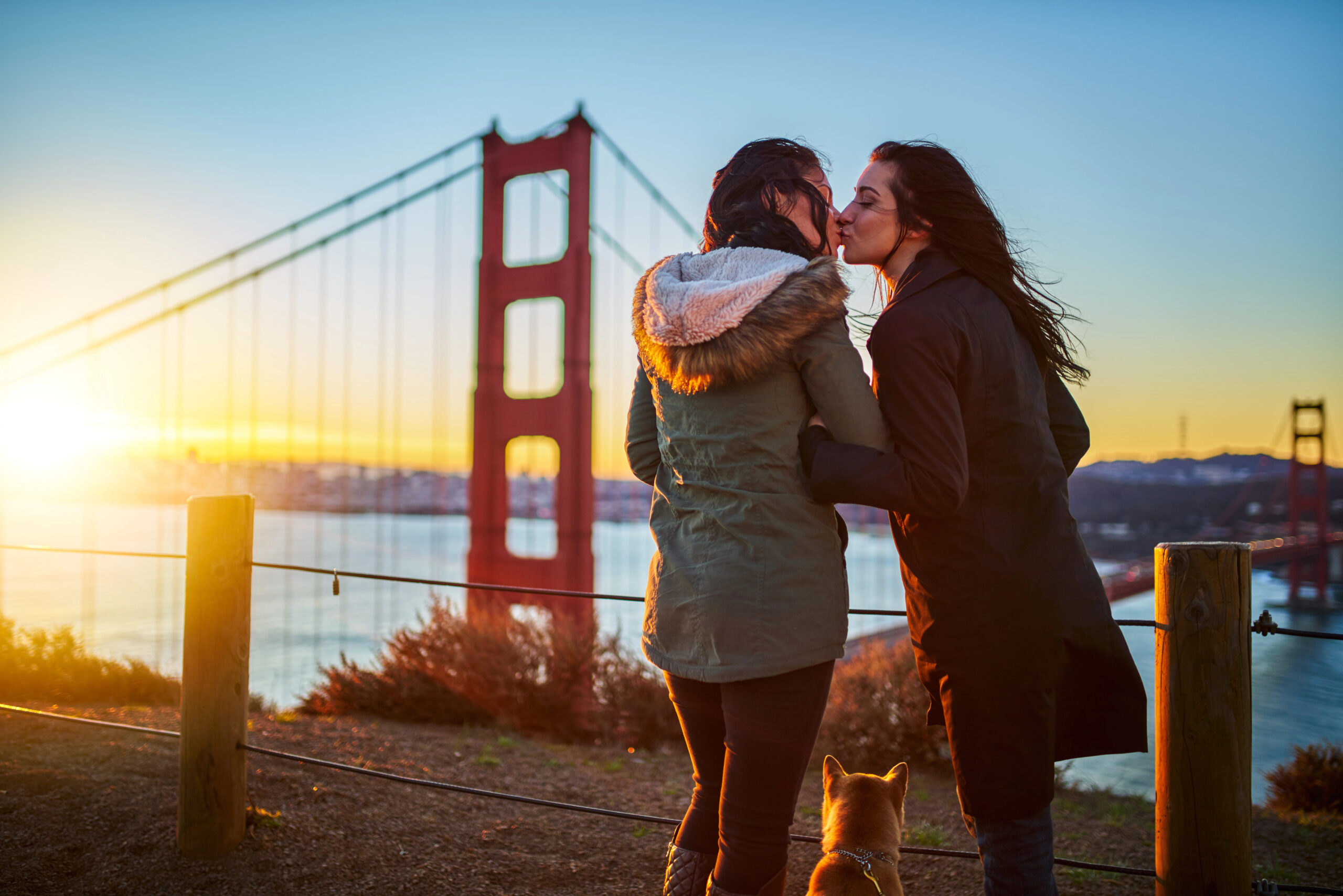 Female couple kissing in front of the Golden Gate Bridge at sunset.