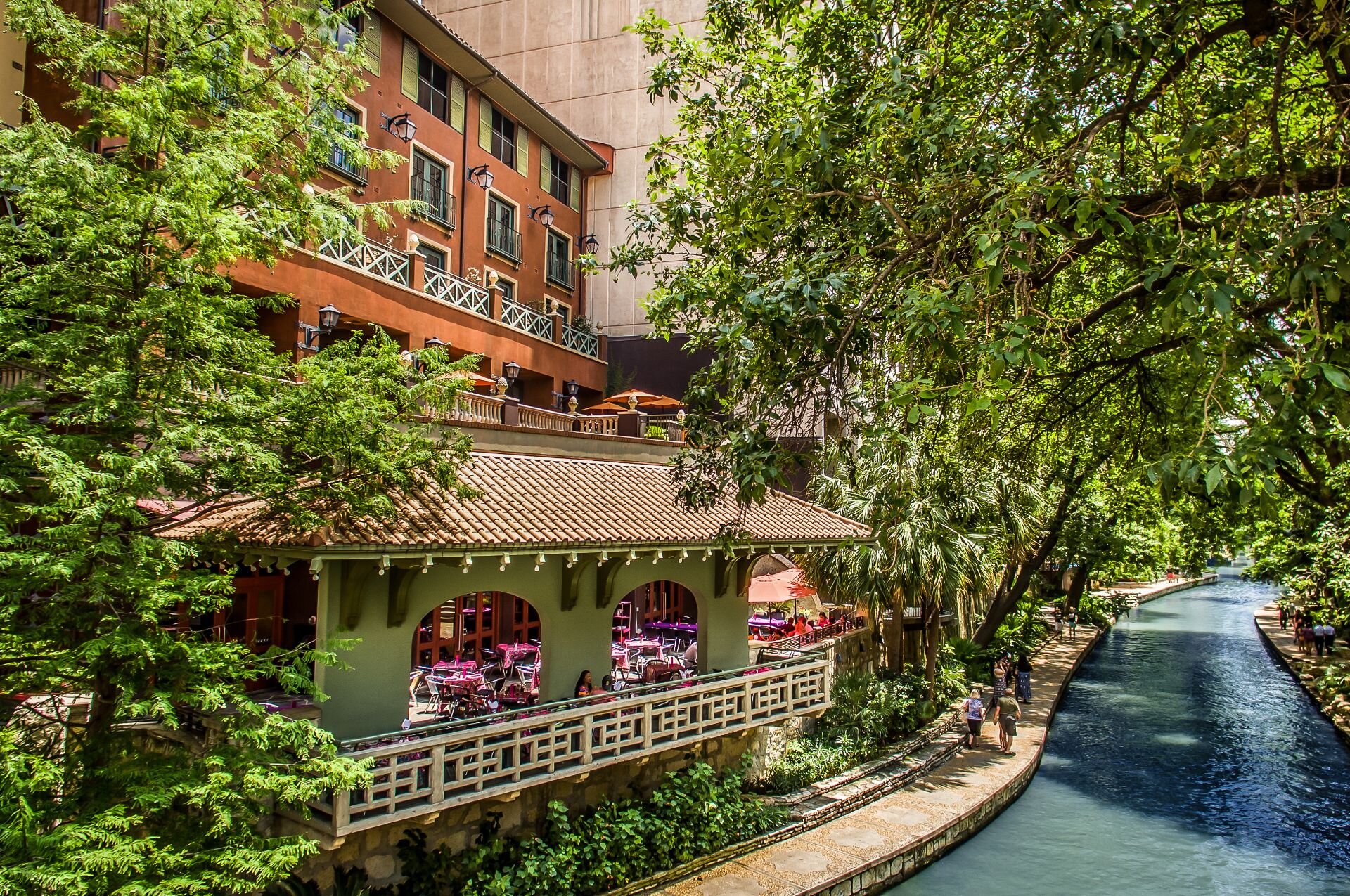 A riverside building features an outdoor dining area and large trees beside a canal on a sunny day. It's one of the most romantic getaways in December.