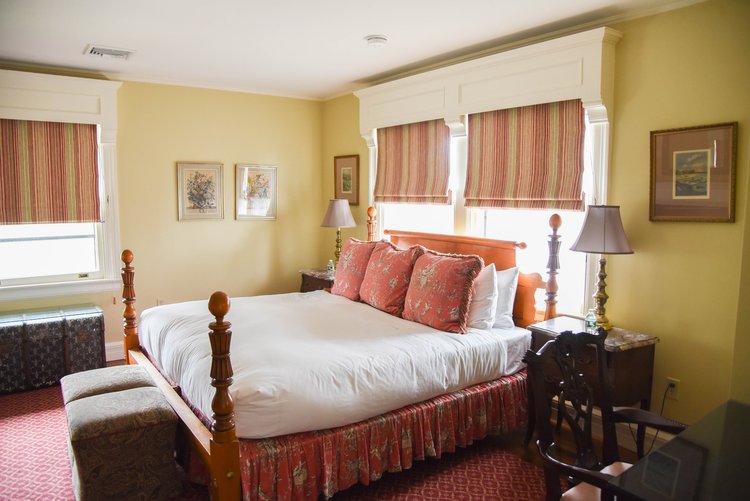 A cozy bedroom includes a large bed with red and white bedding, two bedside tables with lamps, striped window shades, and framed art on the walls.