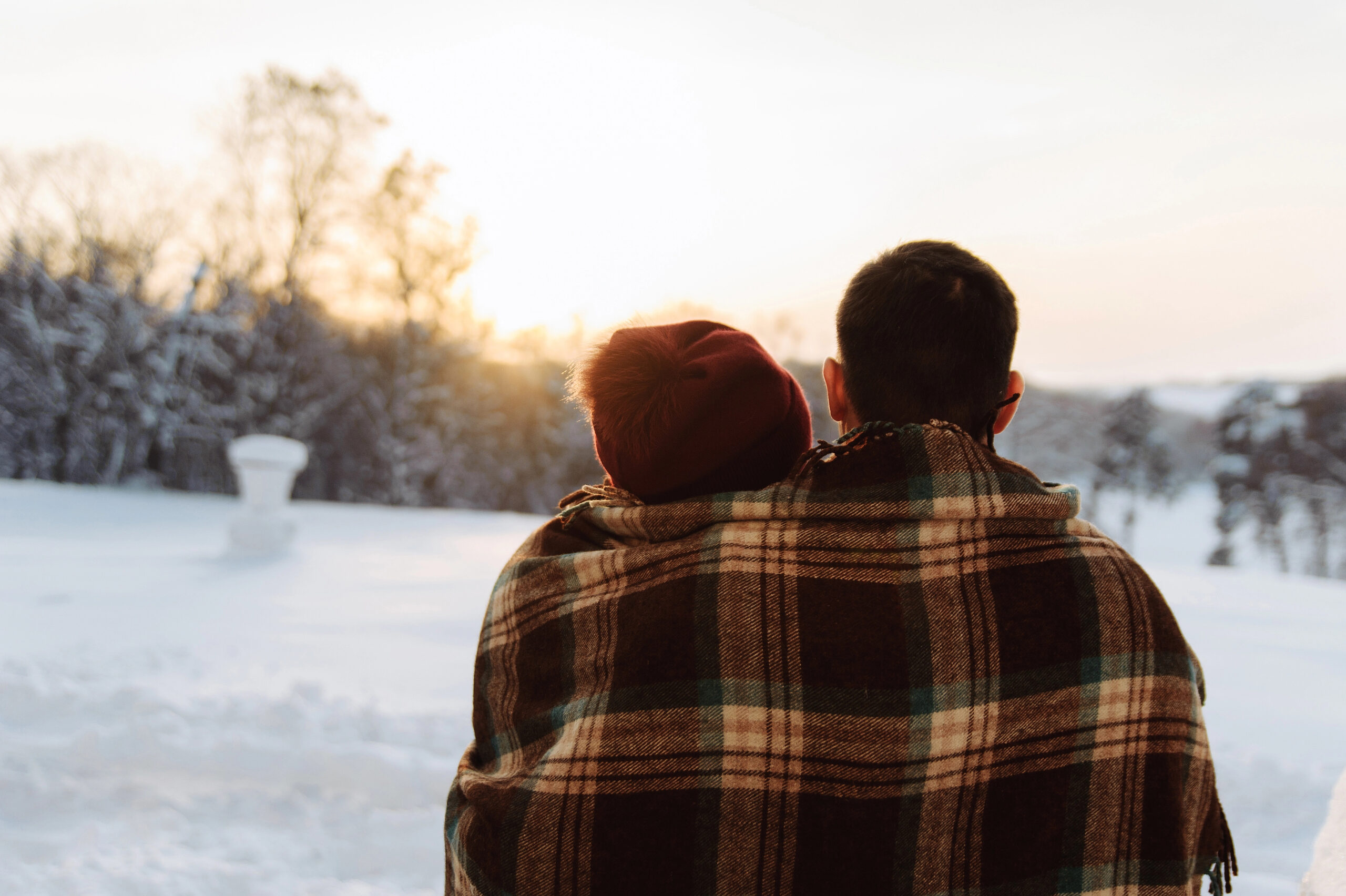 Wrapped in a plaid blanket, two people sit in the snow facing a sunset. It's one of the most romantic getaways in December!