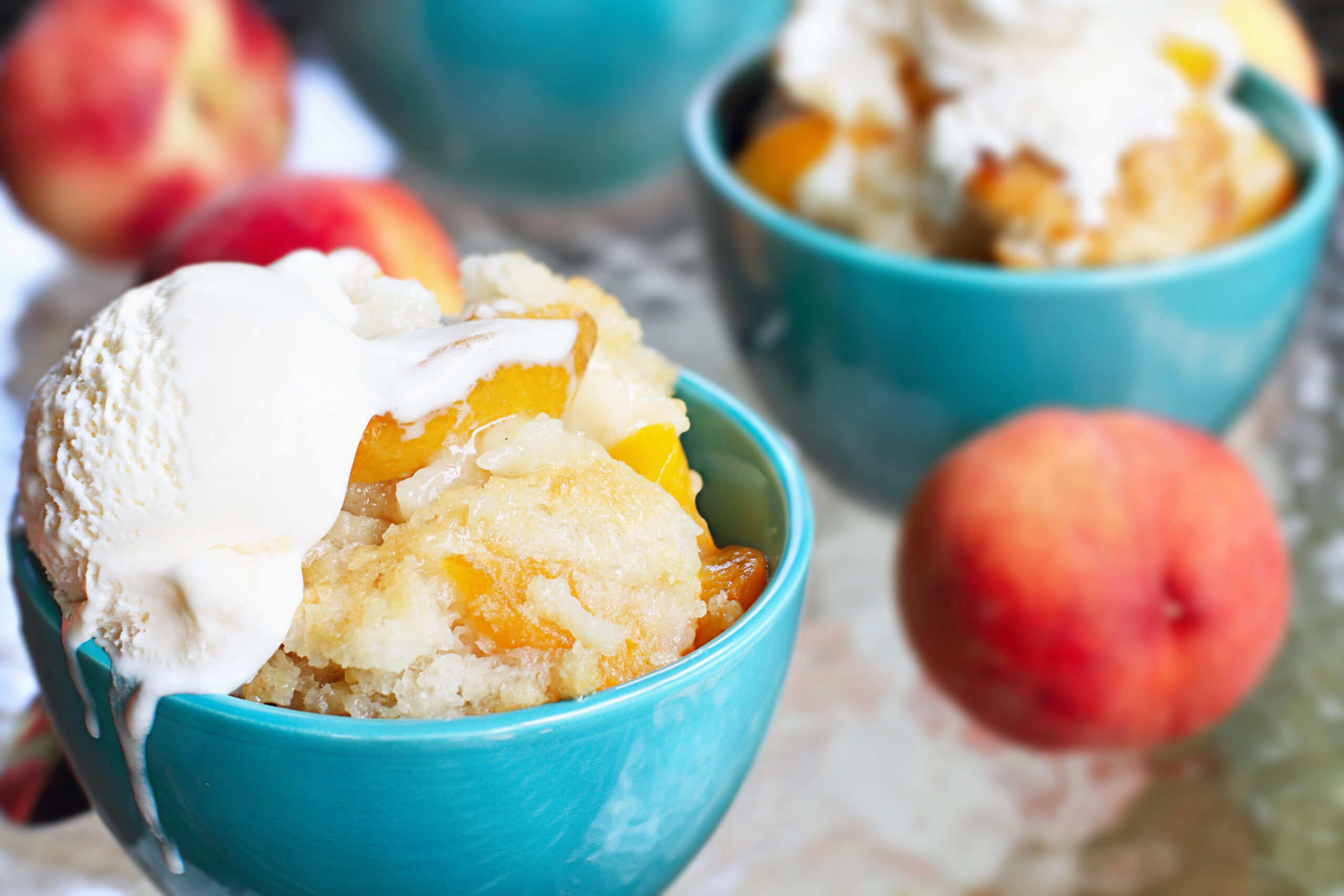 Peach cobbler in a turquoise bowl with ice cream on top and peaches in the background.