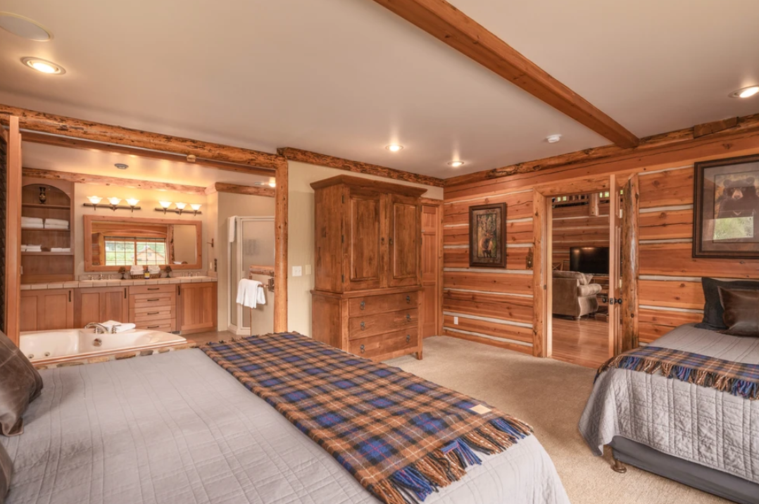 The cozy log cabin bedroom features two beds with plaid blankets, wooden furniture, an open doorway to a bathroom with a tub and sink, warm lighting, and rustic decor.