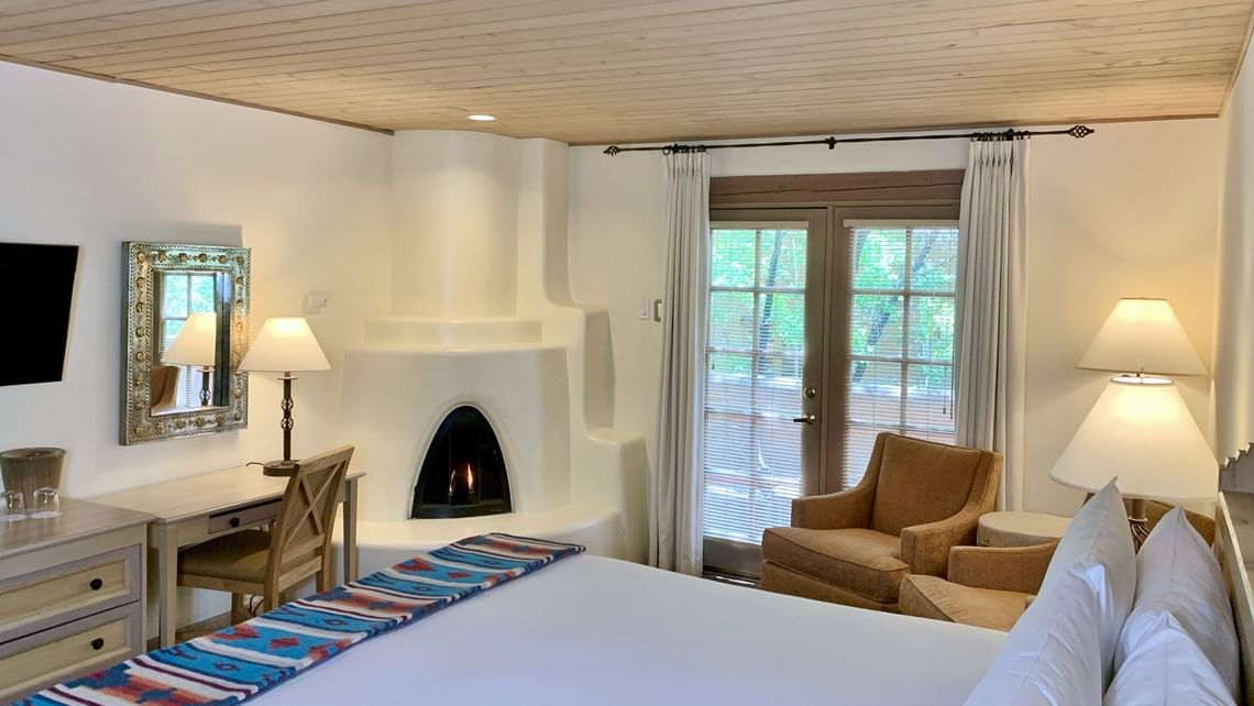 The cozy bedroom features a double bed with soft white bedding, a small fireplace, armchairs, a desk, and a window with curtains allowing natural light. It's one of the most romantic getaways in December!