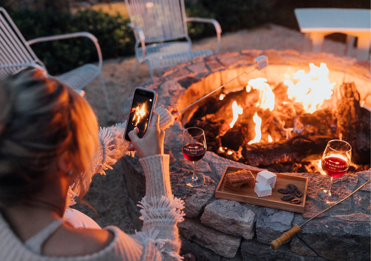 A woman photographs a fire pit surrounded by two glasses of wine, a wooden tray with crackers and chocolate, and roasting marshmallows. It's one of the most romantic getaways in Decembe!