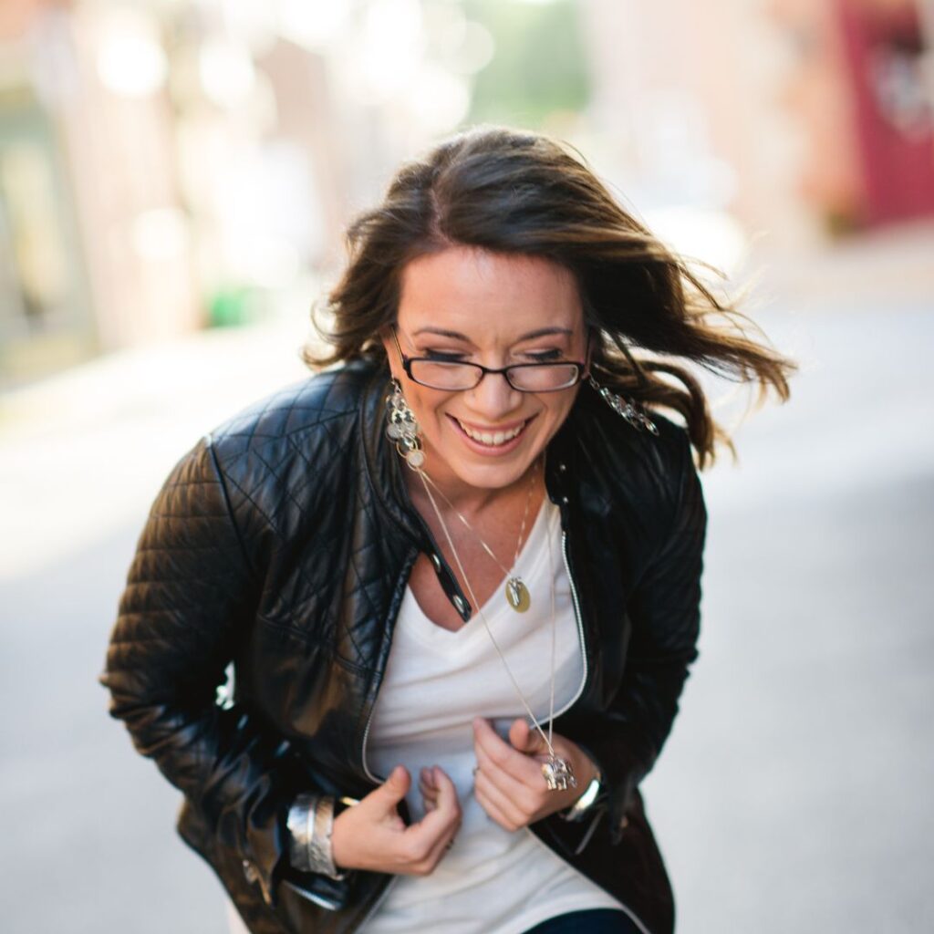 A woman with long brown hair, glasses, a black leather jacket, and a white top smiles and leans forward outdoors.