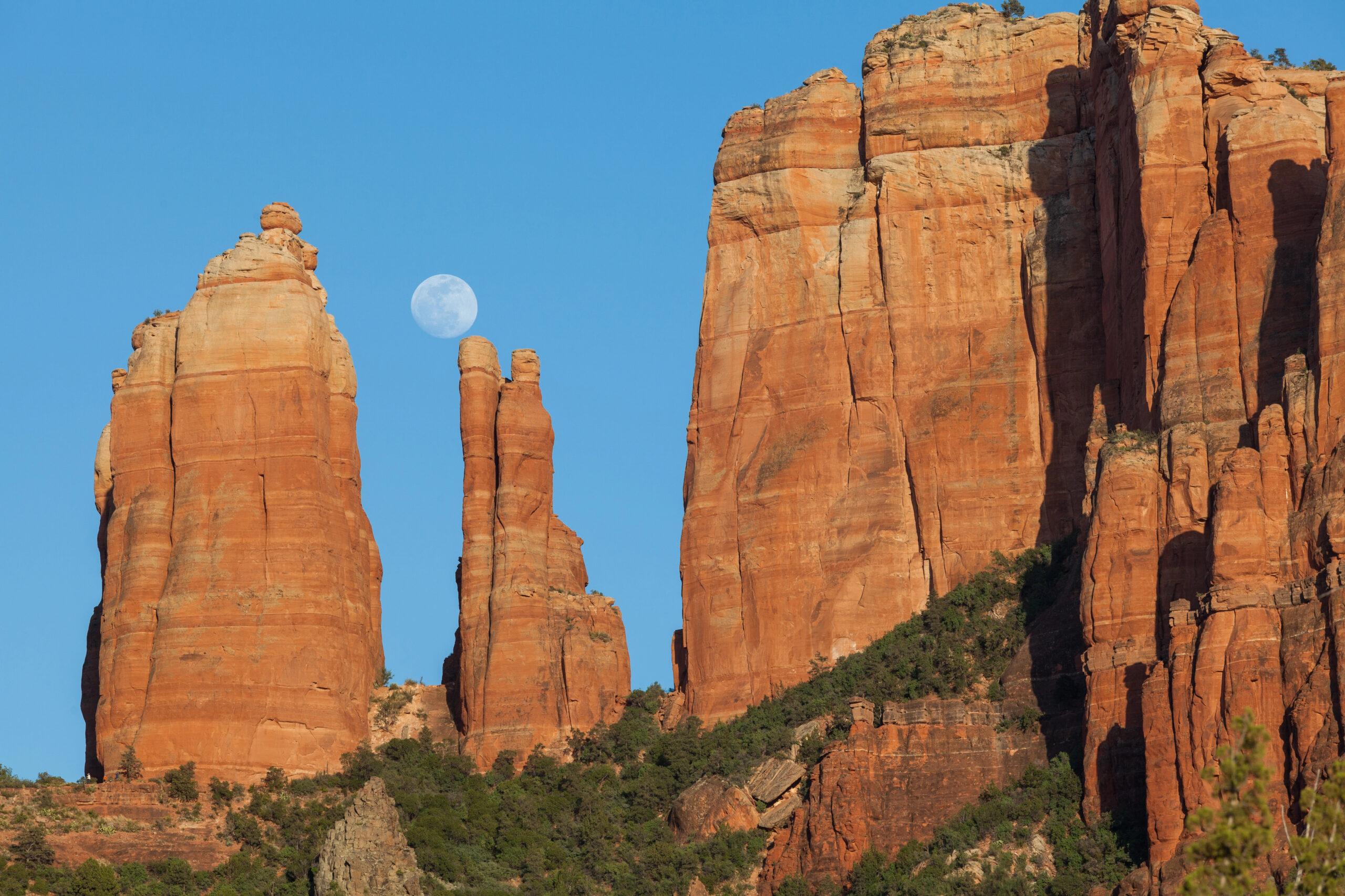 Red rock formations rise under a clear blue sky with the full moon between two spires, while shrubs and trees cover the lower slopes.