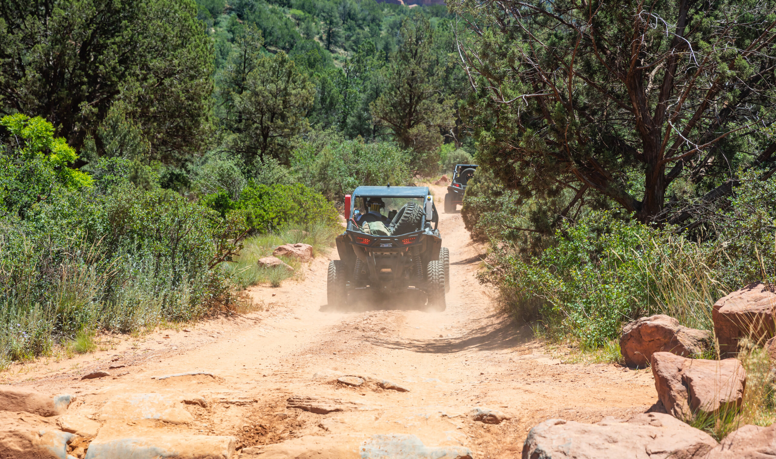 Two off-road vehicles travel a dusty dirt trail through a forested area in Sedona, flanked by trees and shrubs.