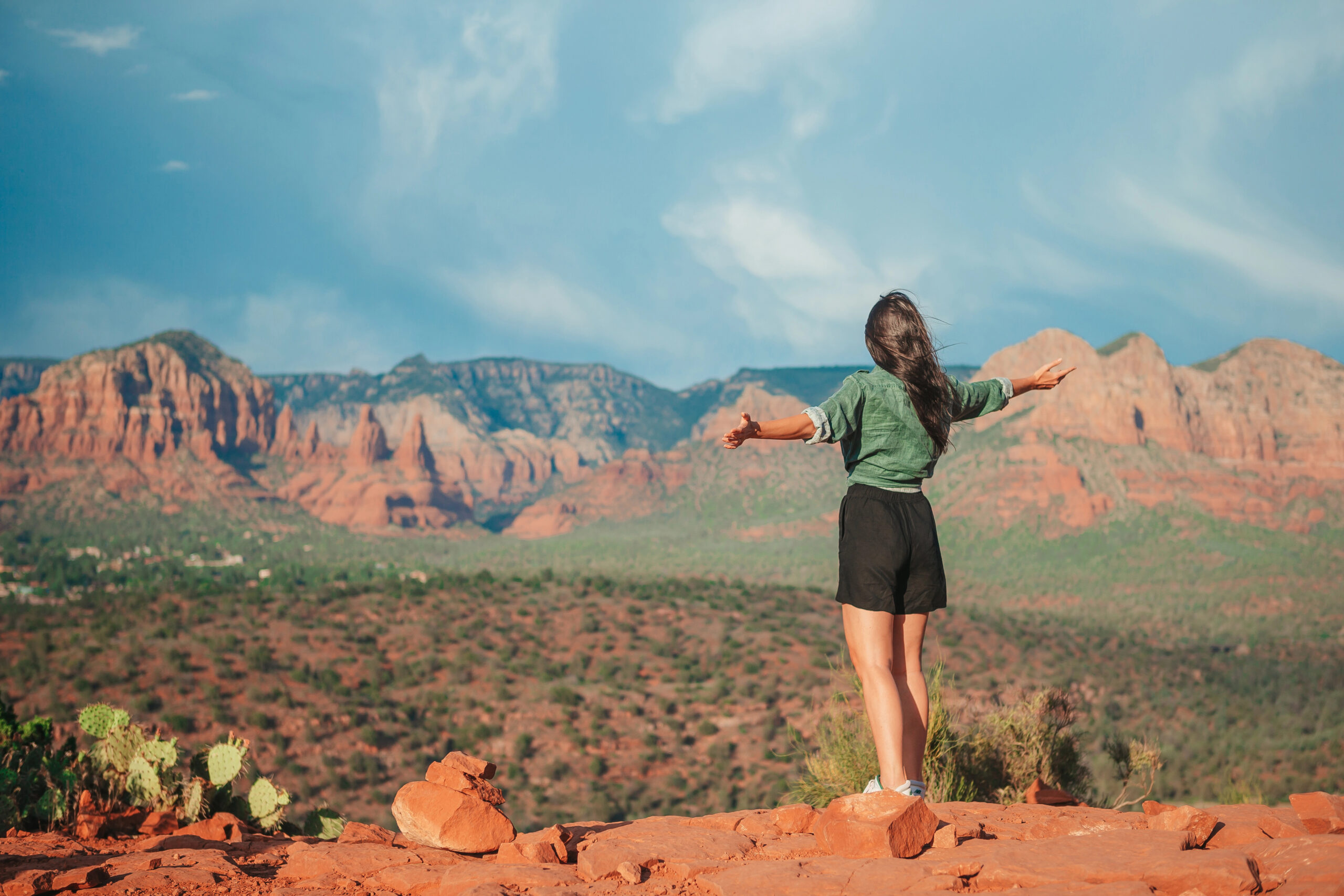 A person stands on red rocks with arms outstretched, facing desert mountains beneath a blue sky.