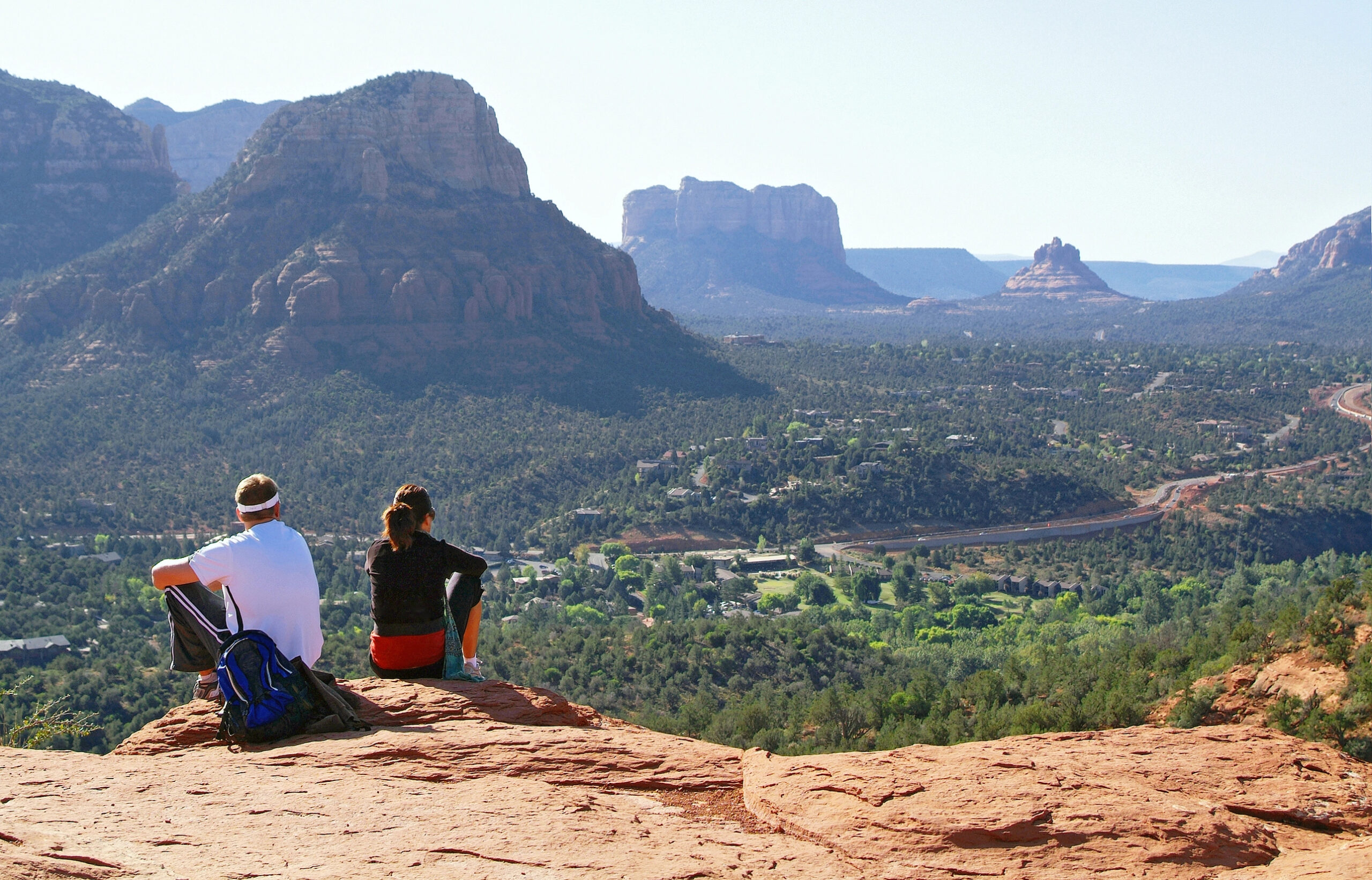 A couple enjoying a weekend in Sedona, sitting on a rocky ledge overlooking a scenic valley of mountains.
