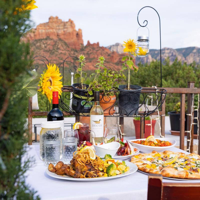 Outdoor dining table set with dishes, wine, and glasses, surrounded by potted plants and sunflowers, overlooking Sedona’s red rock mountains.