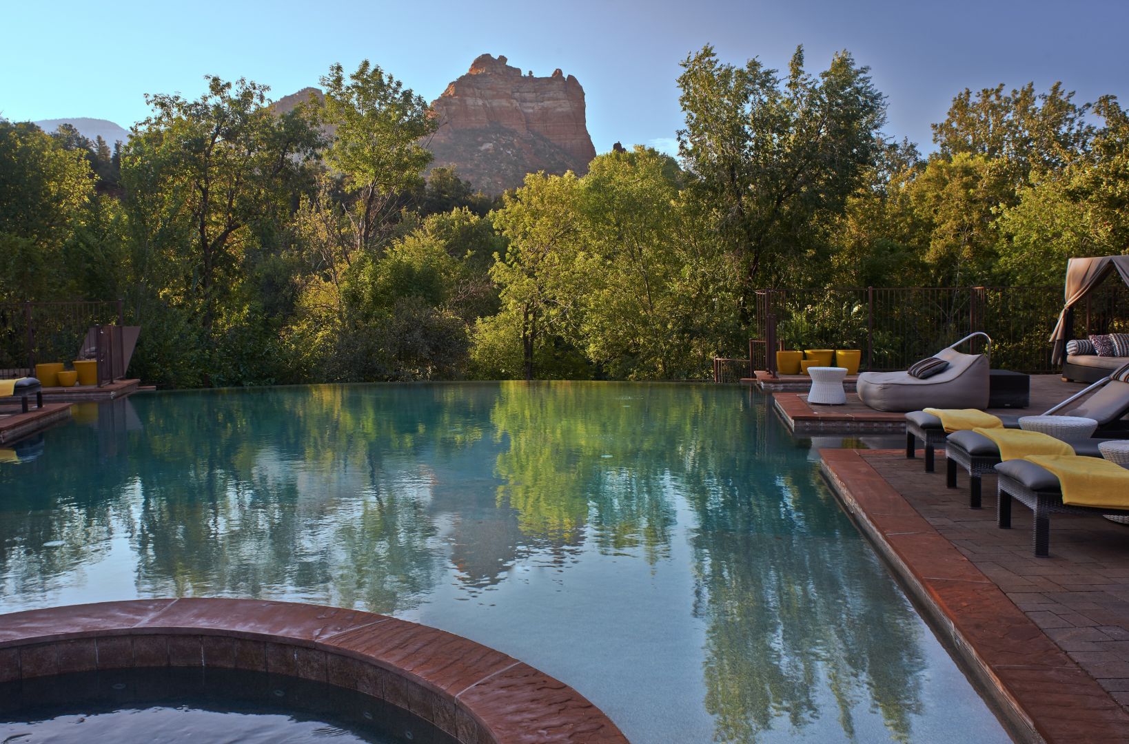 Infinity pool, lounge chairs, and a hot tub surrounded by trees with a rocky mountain backdrop under a clear sky.