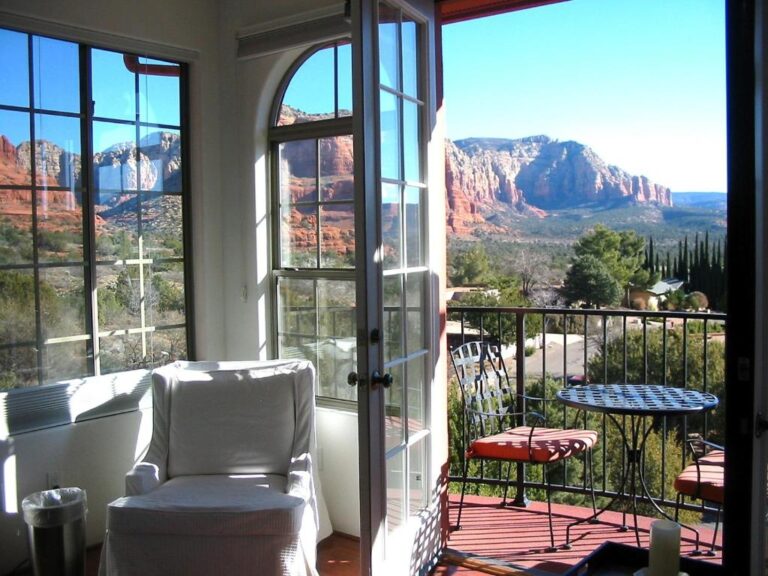 A bright room with a white armchair features open glass doors to a balcony with seating and views of red rock mountains beneath a clear blue sky.