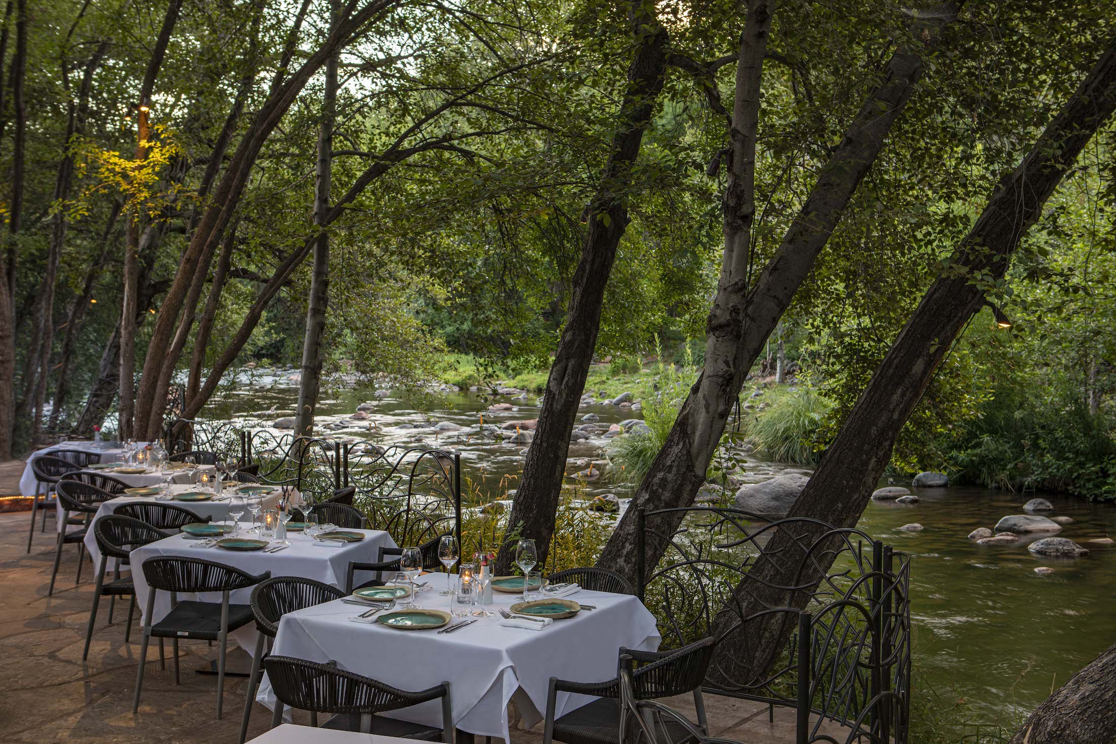 Outdoor restaurant tables with white tablecloths and place settings are set beside a tree-lined, rocky river surrounded by lush greenery.