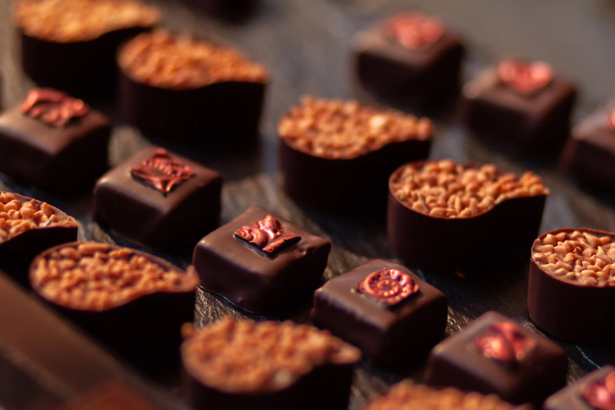 Assorted dark chocolates with textured toppings and embossed designs are arranged on a dark surface.