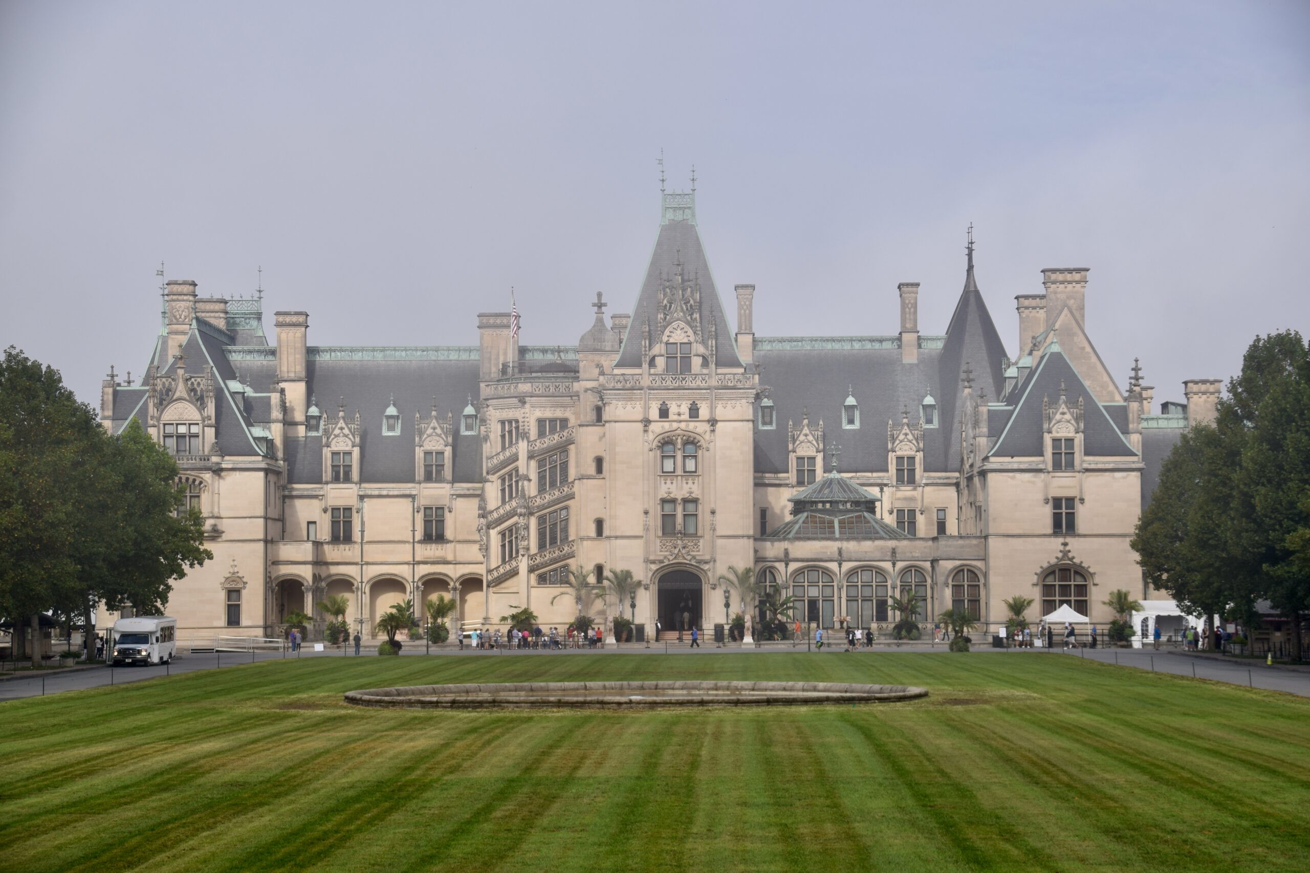 A historic mansion with steep roofs and many windows stands behind a circular fountain and striped lawn, framed by trees under a foggy sky.