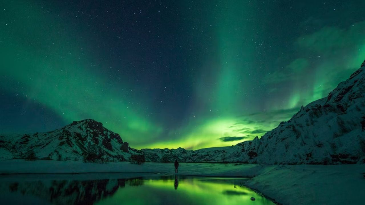 A lone figure stands by a reflective lake beneath vivid northern lights in snowy mountains, captured in Norway.