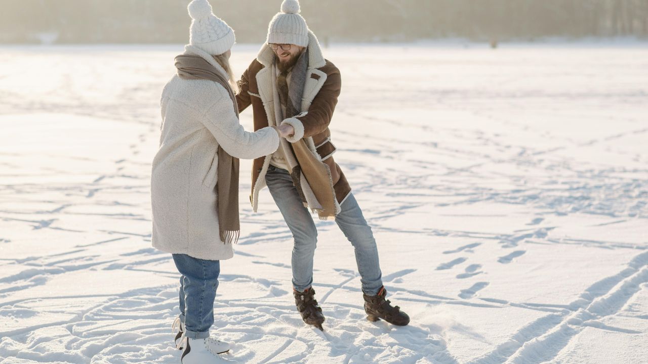 Happy couple ice skating together on a snowy frozen lake in winter, wearing cozy coats and knit hats
