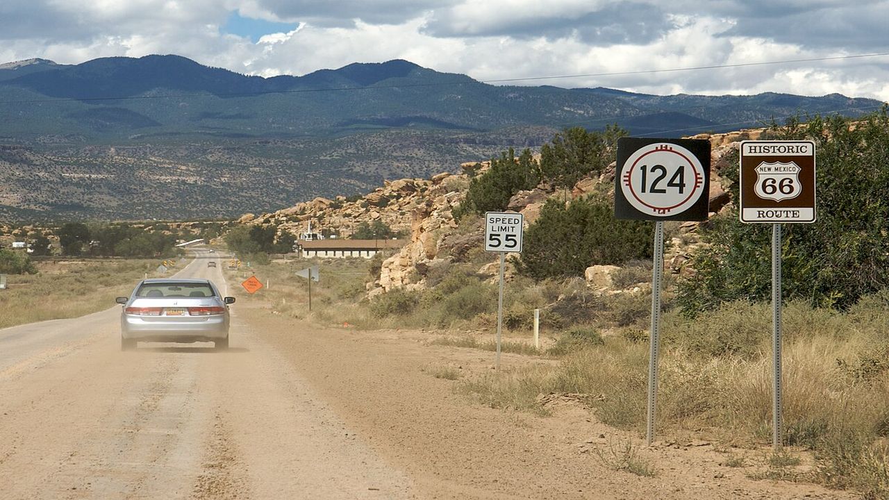 A car drives along a dusty, open road with rugged hills and mountains in the background. Road signs and desert vegetation line the way.