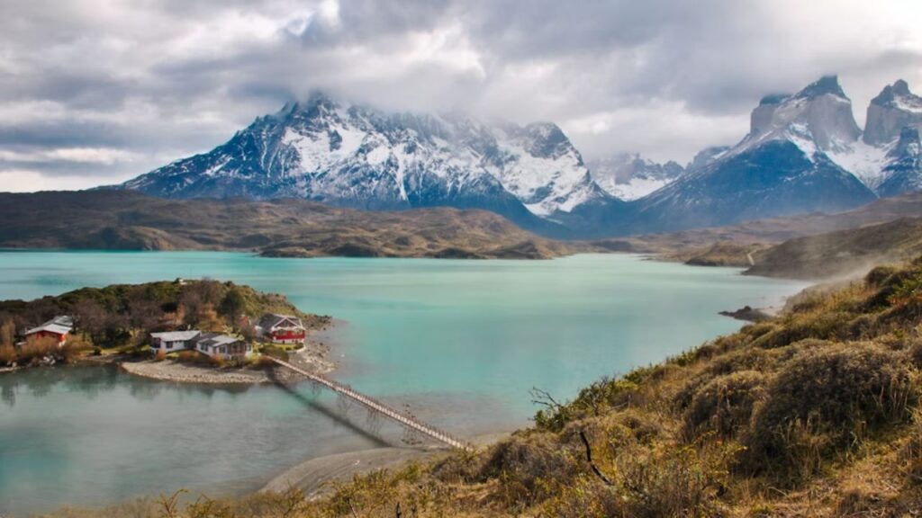 Turquoise lake and snow-capped peaks in Torres del Paine National Park, Chile, with cozy lodges on a small island connected by a footbridge.