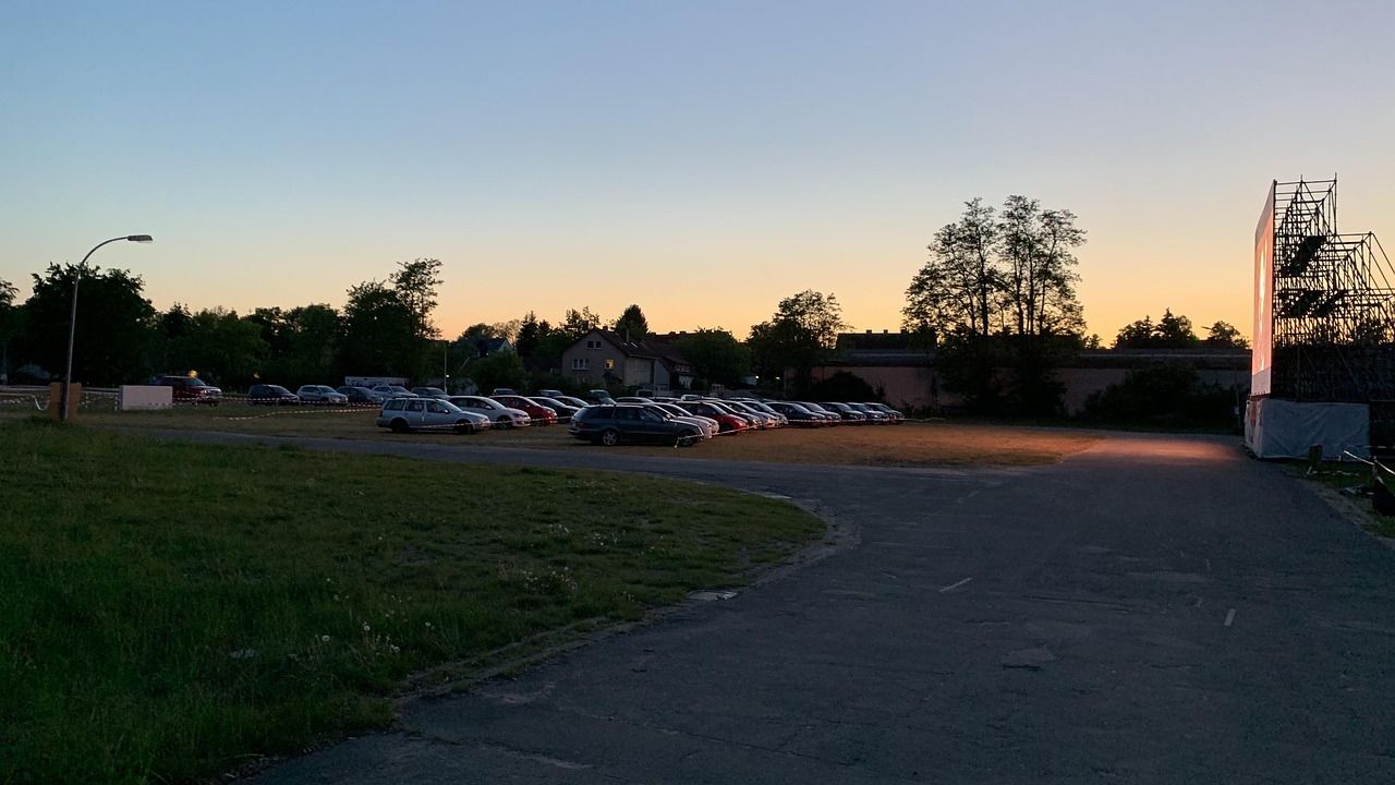 Drive-in theatre at dusk with parked cars facing a lit screen and a sunset sky in the background.