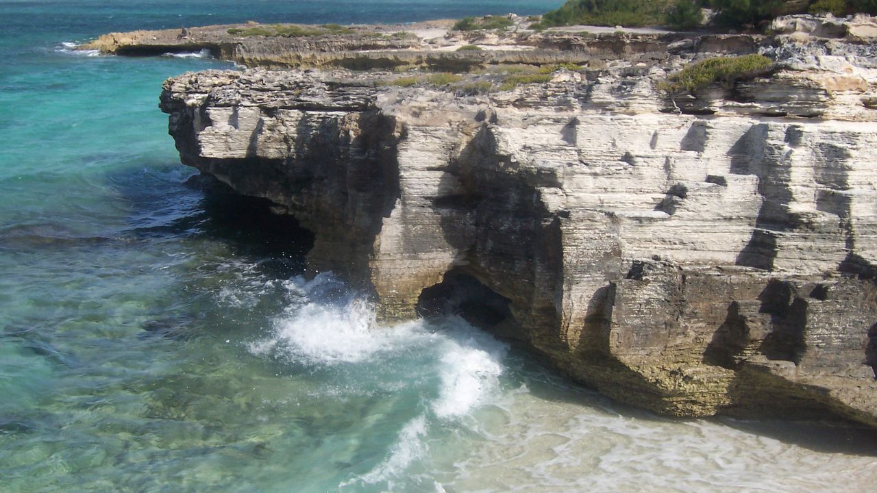 Waves crash against rugged limestone cliffs, forming small sea caves at the base. The turquoise water contrasts with the pale, weathered rock layers.