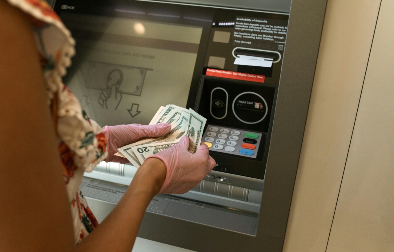 Person in gloves holding dollar bills at an ATM.
