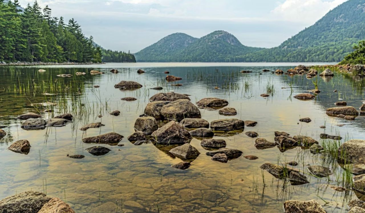A calm lake with rocky shallows is surrounded by green forests and hills beneath a partly cloudy sky.