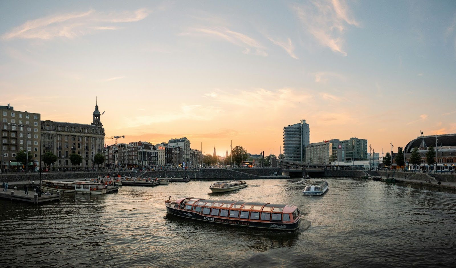 Tour boats and smaller vessels navigate a city river at sunset, with buildings and a bridge in the background.