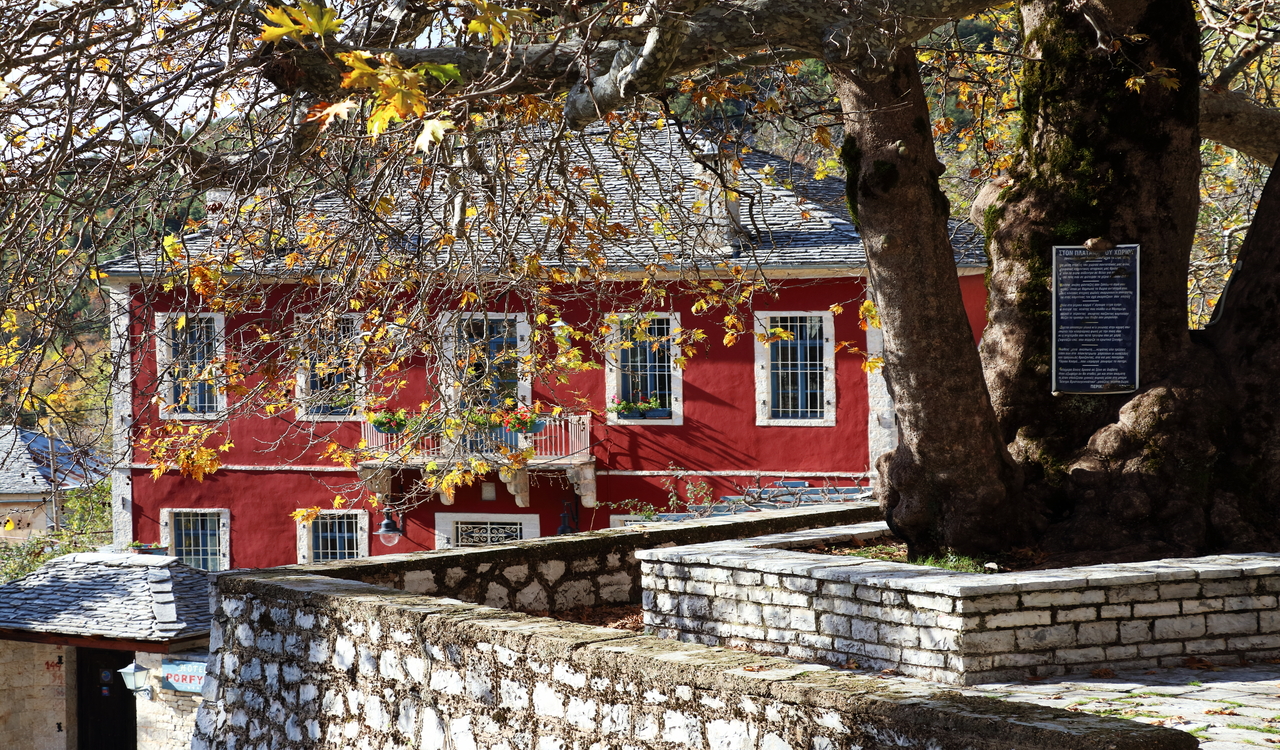 Historic red stone house in Ano Pedina, Greece, framed by autumn trees and a centuries-old courtyard.