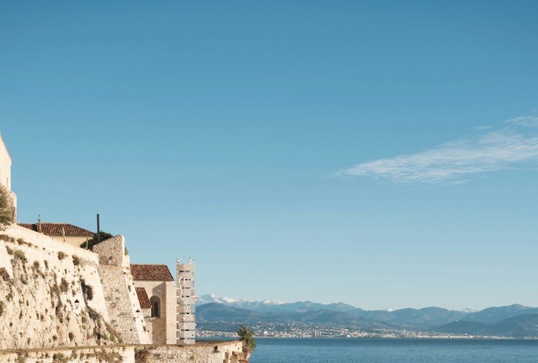 Stone buildings perch on rocky cliffs above the sea, with distant mountains beneath a clear blue sky.
