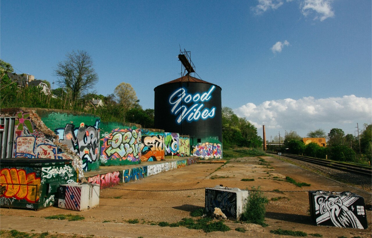 A large water tower reading “Good Vibes” in neon stands amid colorful graffiti by railroad tracks under a partly cloudy sky.