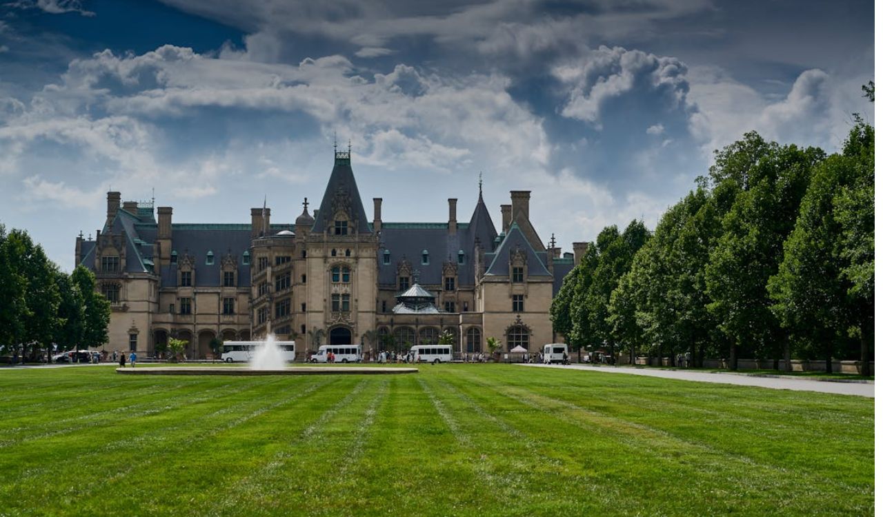 A large historic mansion with steep roofs and turrets stands amid green lawns and trees under a partly cloudy sky, with a fountain in front.