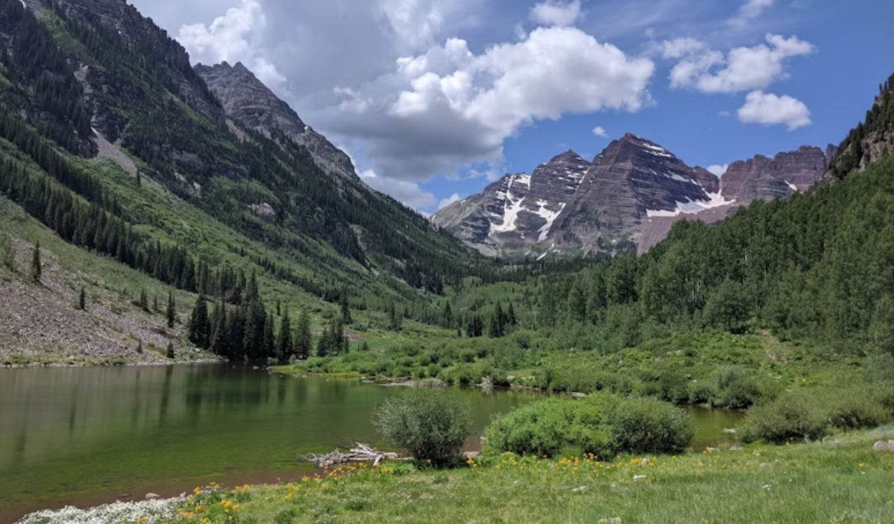 A clear lake lies in a green valley bordered by forested hills and snow-capped mountains beneath a partly cloudy sky.