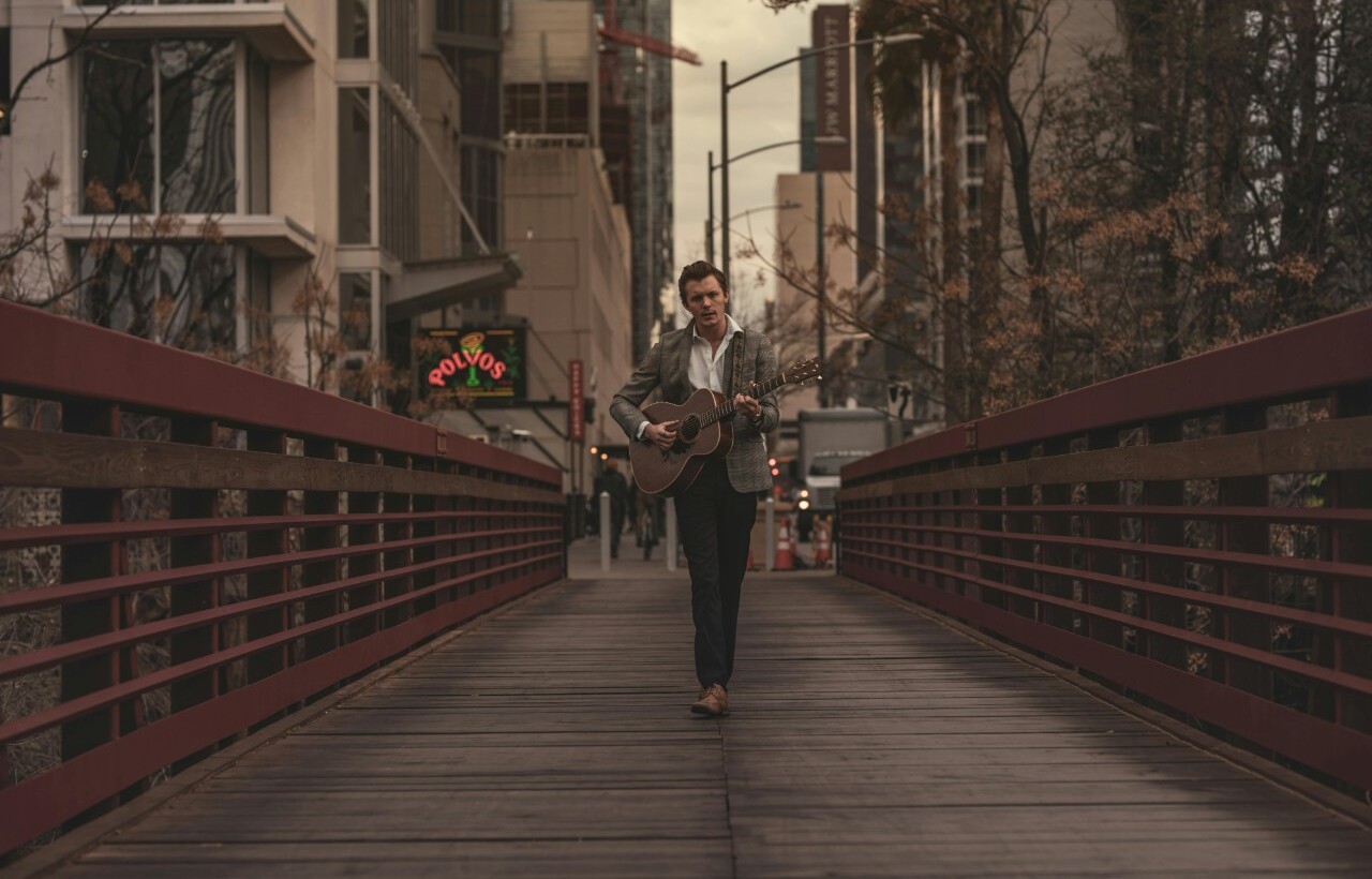 A man in a suit plays an acoustic guitar while walking on a wooden bridge in an urban area with buildings and a neon sign in the background.