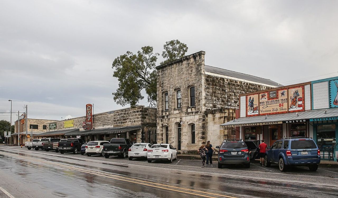 Historic stone and colorful storefronts line a wet street with parked cars and pedestrians under a cloudy sky.