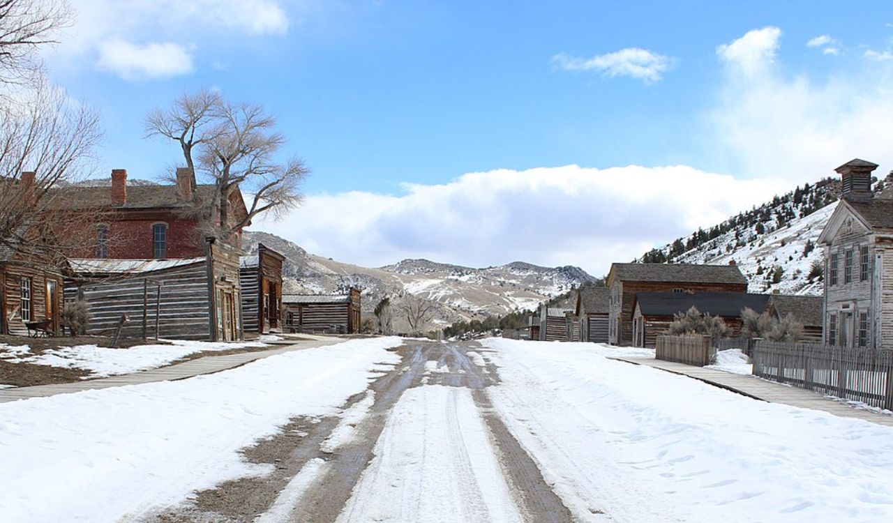 A snow-covered street runs through a deserted old town of wooden buildings, with hills in the background beneath a partly cloudy sky.