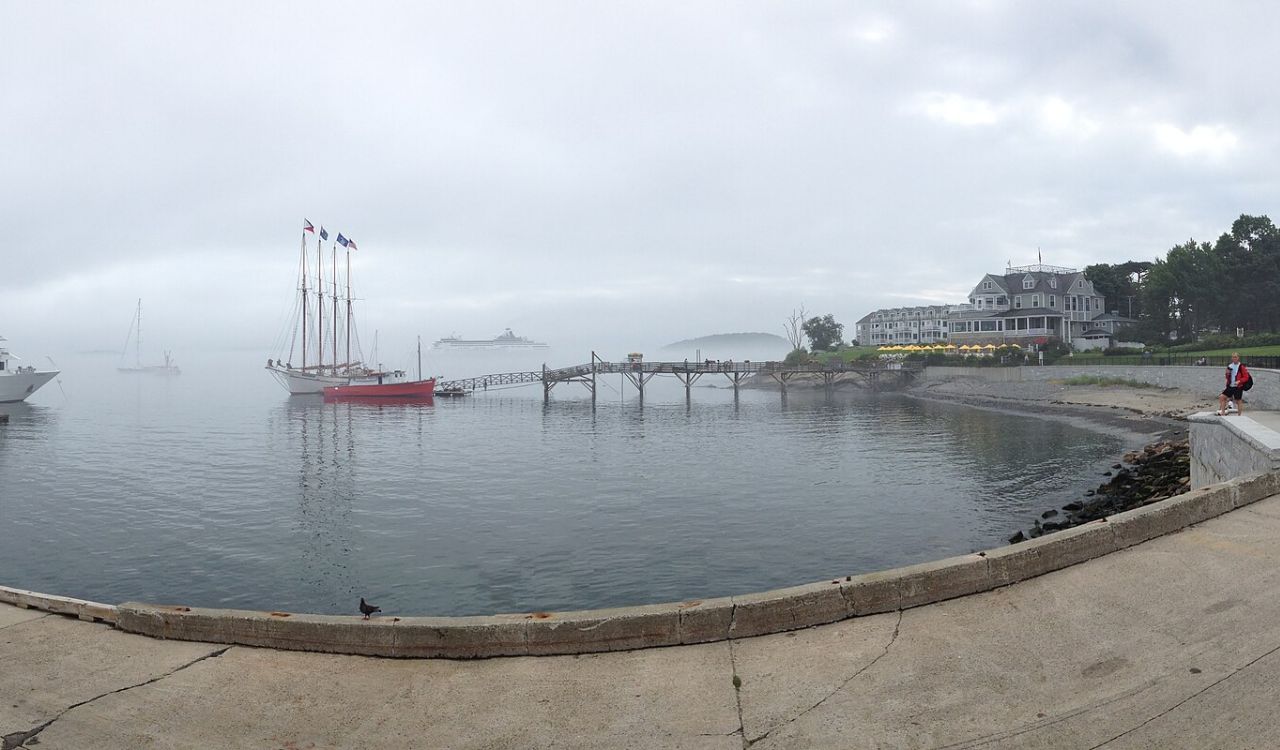 A foggy waterfront features sailboats, a wooden pier, a large building with a porch, and someone sitting on a concrete shore.