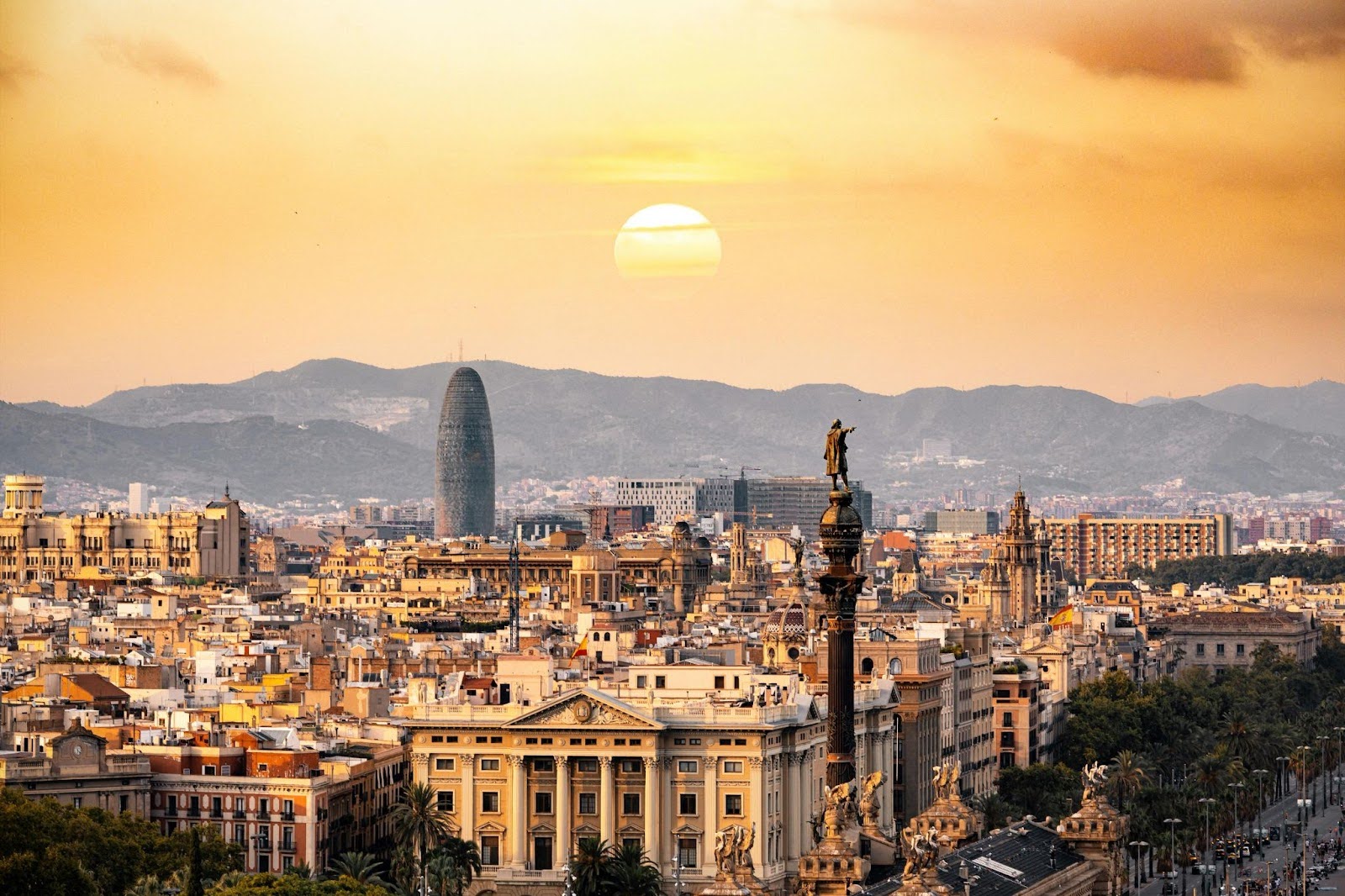 A panoramic sunset view of Barcelona showcases city buildings, the Columbus Monument, and mountains silhouetted against the setting sun.