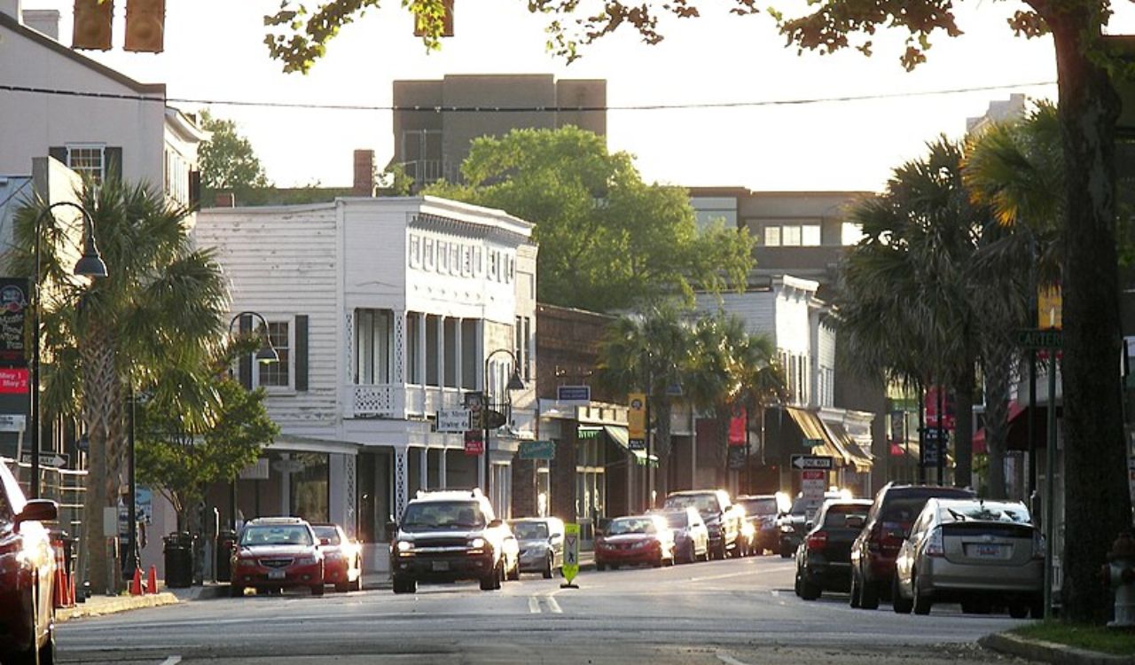 Cars travel down a tree-lined street bordered by shops and buildings in a small town during the day.