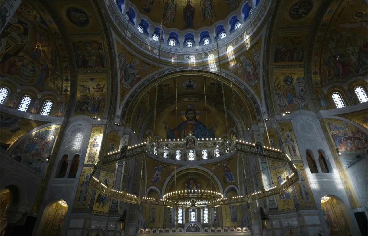 Ornate cathedral interior with large domes, arches, religious iconography, and stained glass windows illuminated by natural light.